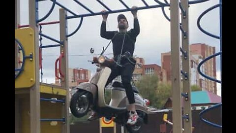 Strong man raises the bar by doing pull ups while attached to motor scooter