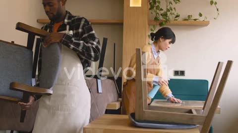 Waitresss Cleaning Coffee Shop Table With Disinfectant Spray And Rag While Waiter Arranging Chairs