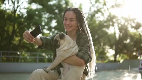 Smiling girl taking selfie photo with cute pug puppy in green city park