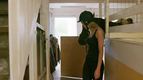 Young female jockey is leading a brown horse away through the stables