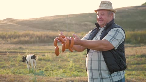 Happy adult farmer smiling at camera holding sausages in hands with herd of cows in cowshed on farm