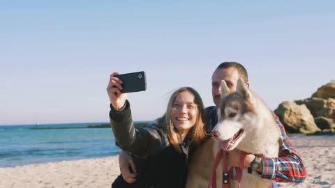 Young caucasian couple on beach doing selfie on smart phone with siberian husky dog