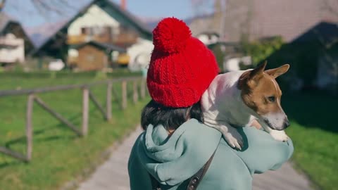 Young girl in red cap carrying a small dog on her shoulder and walking at countryside on grass lawn