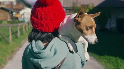 Young girl in red cap carrying a small dog on her shoulder and walking at countryside on grass lawn