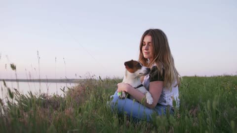 Young happy woman and het little dog sitting with flying kite on a glade at sunset