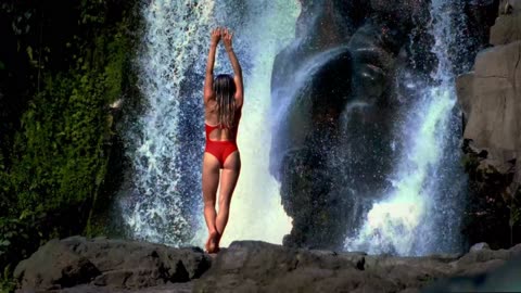 A beautiful woman in a bikini is seen breathing fire in the middle of a beautiful mountain waterfall