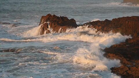 Waves crash against rugged rocks at the shoreline during sunset in a coastal setting