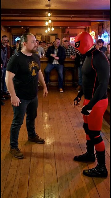 Russell Blade bodyslams a red mask wrestler on the wooden floor at a bar.