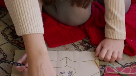 A little girl positioned comfortably on the floor receives help from an adult.