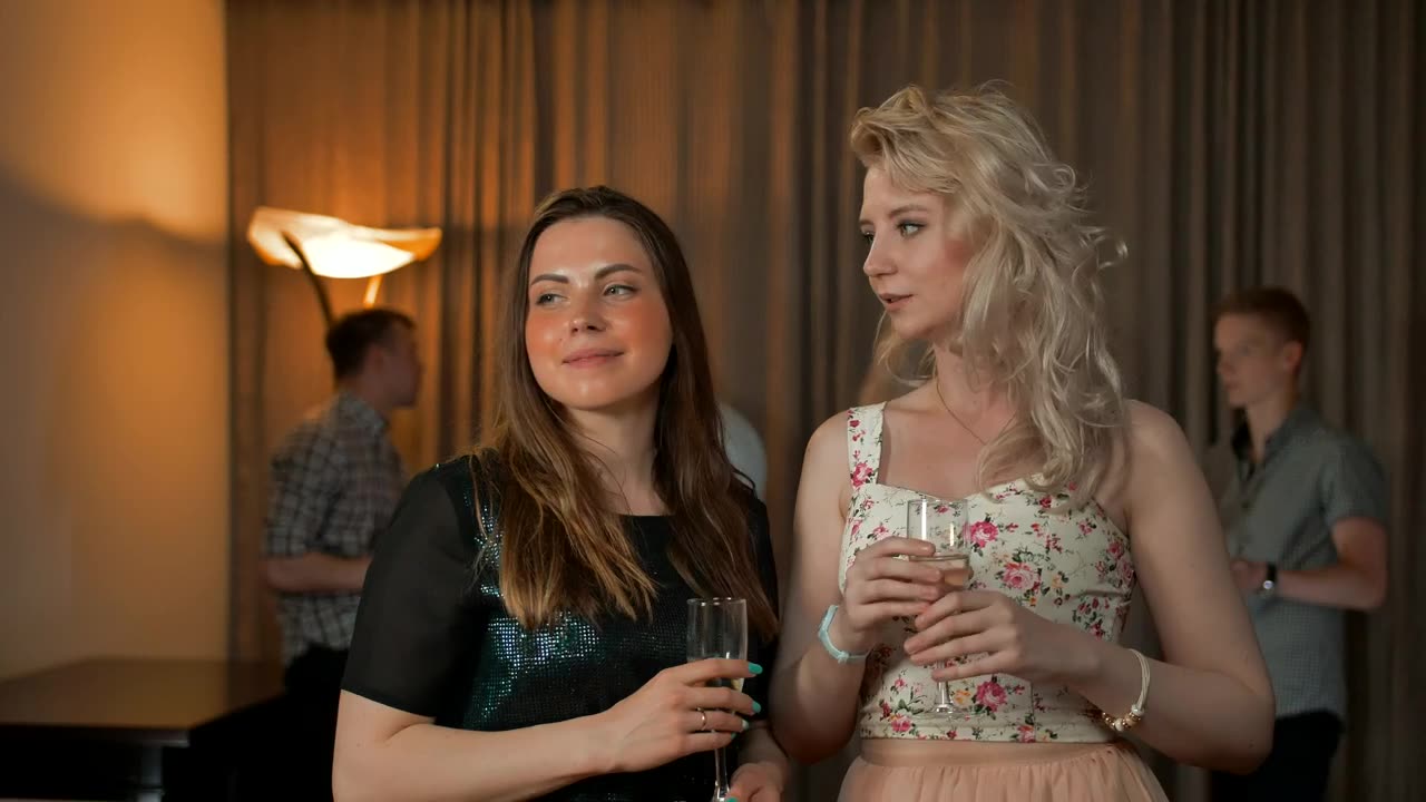 A couple of girls at a party drinking champagne while gossiping in each other's ears.