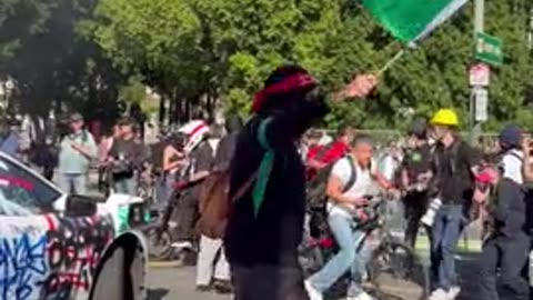 Protester waves Mexican flag in front of burning WayMo vehicle.