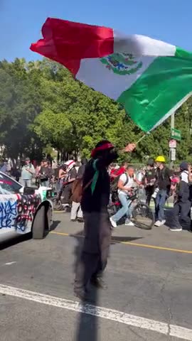 Protester waves Mexican flag in front of burning WayMo vehicle.
