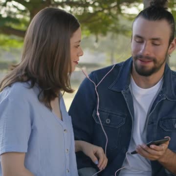 A young happy couple share a wired heaphone at the park bench