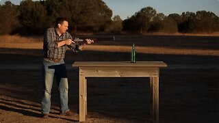 A Man with a Shot gun shooting a Glass Bottle from a table