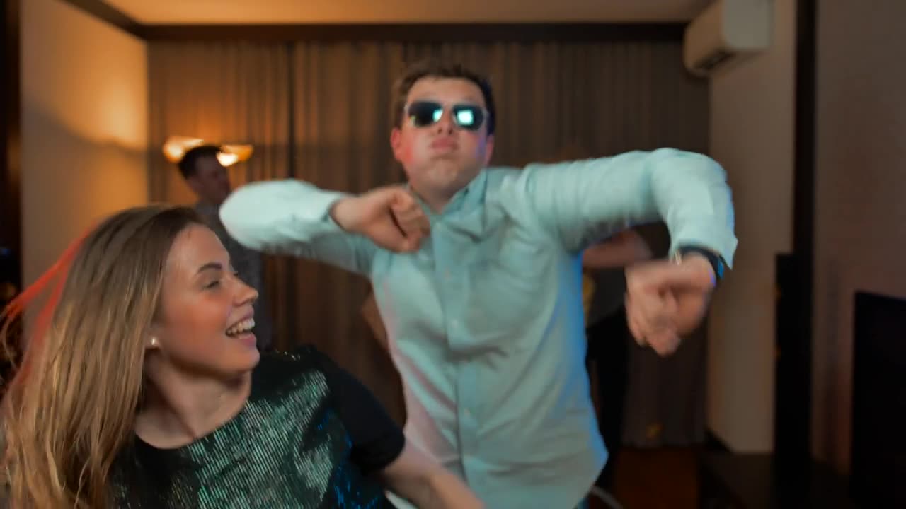 A young couple dancing in front of the camera as another group behind, inside a living room.