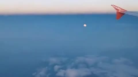 A passenger on a flight records the moon appearing among the clouds below the airplane