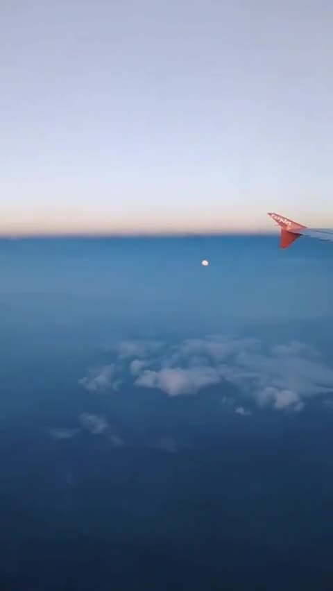A passenger on a flight records the moon appearing among the clouds below the airplane