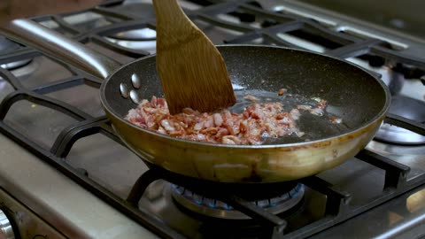 Close-up shot of a stove while a person frying diced bacon in a skillet.