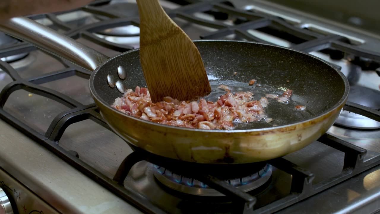 Close-up shot of a stove while a person frying diced bacon in a skillet.