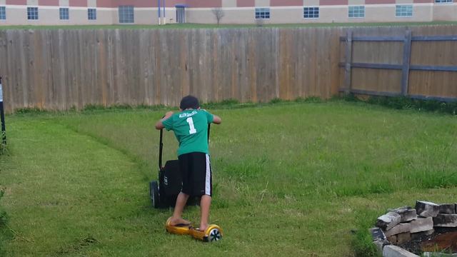 A Boy Rides A Hoverboard While He Mows The Lawn With A Push Mower