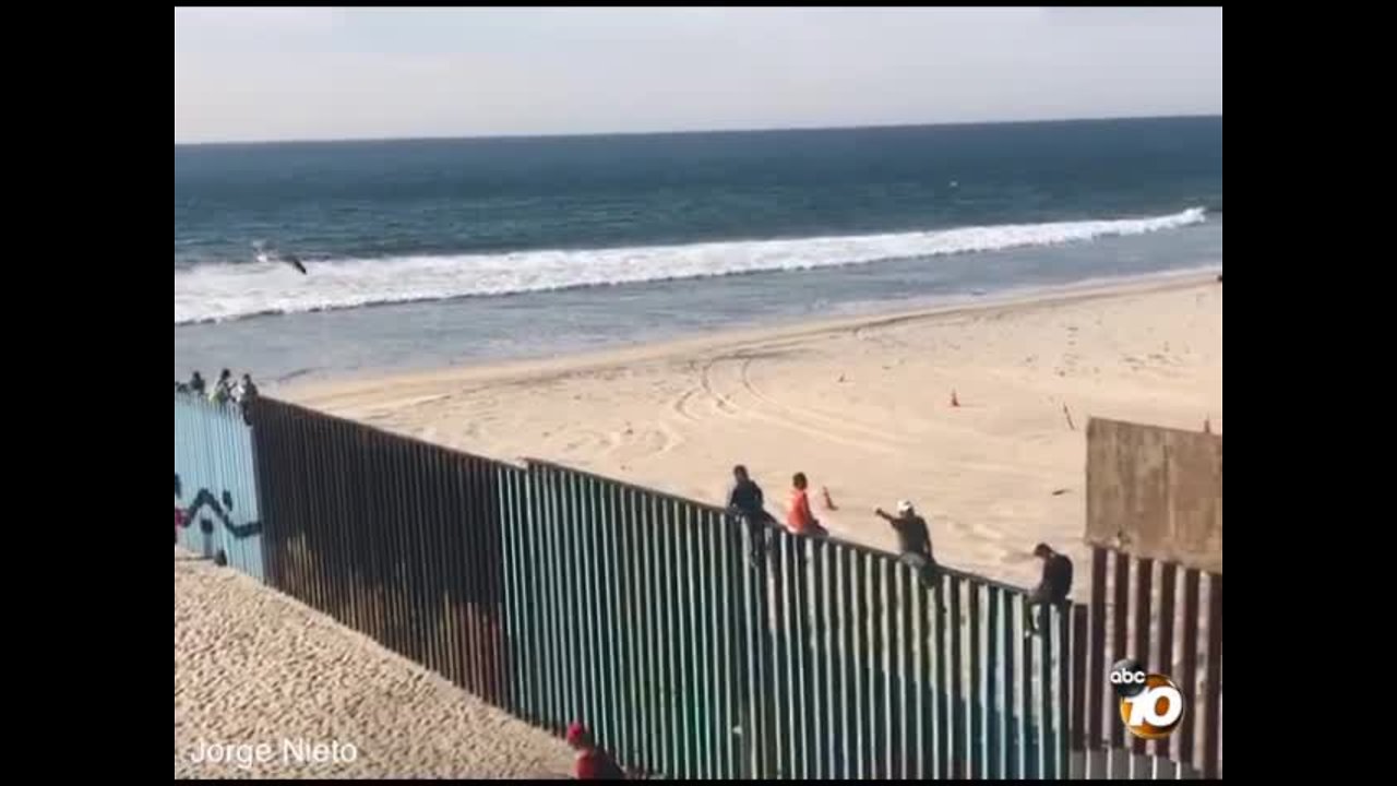 People climb on top of border fence as caravan approaches