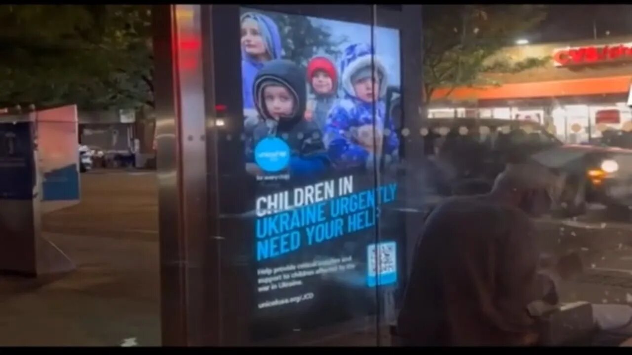 A homeless New Yorker’s dinner is illuminated by “CHILDREN IN UKRAINE NEED YOUR HELP” bus stop sign