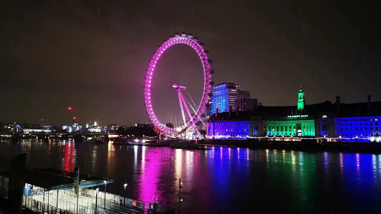 A rainy English night at the London eye, with a colourful light display in the background