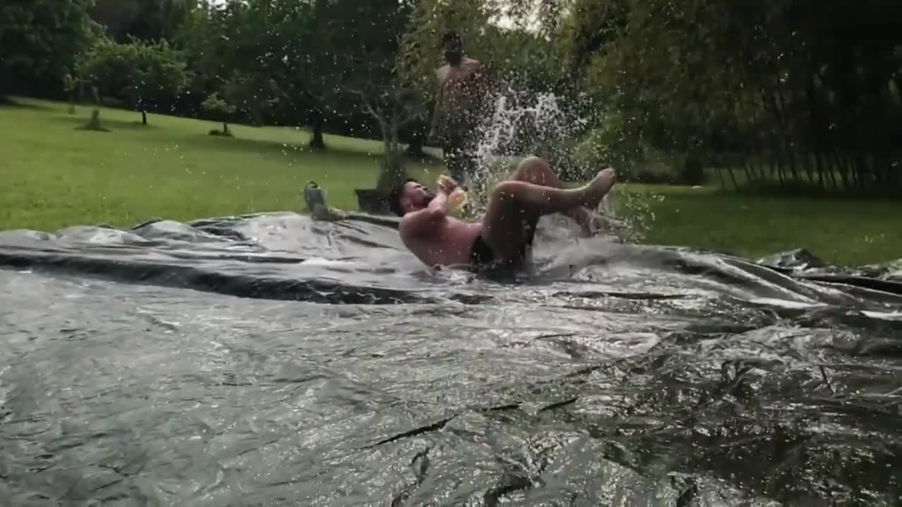 Guy Drinking Beverage Slides Over Massive Sheet of Plastic on Grass