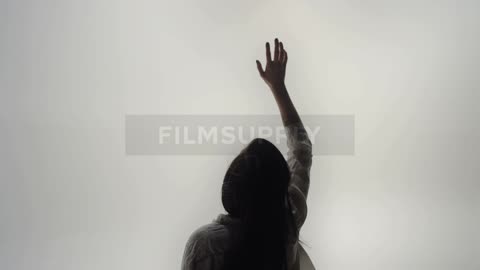 Woman with straight hair and white shirt raises her hand against the cloudy sky