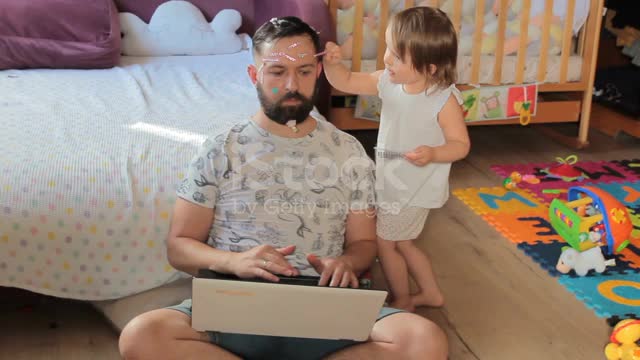 Dad working from home. His little daughter sticks shiny stickers on his face.