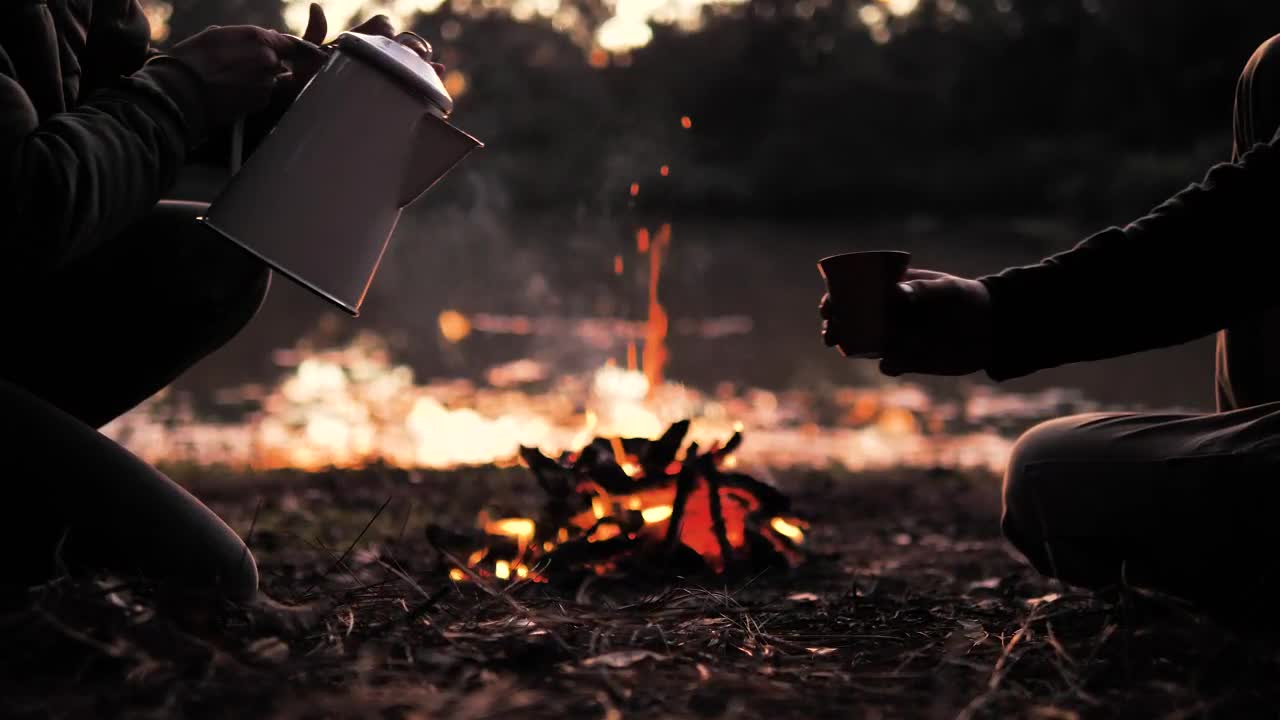 People sharing a warm drink around a campfire in the early evening.