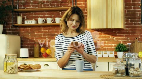 Young Attractive Woman Sitting At The Kitchen With A Coffee In The Morning And Tapping Or Typing On