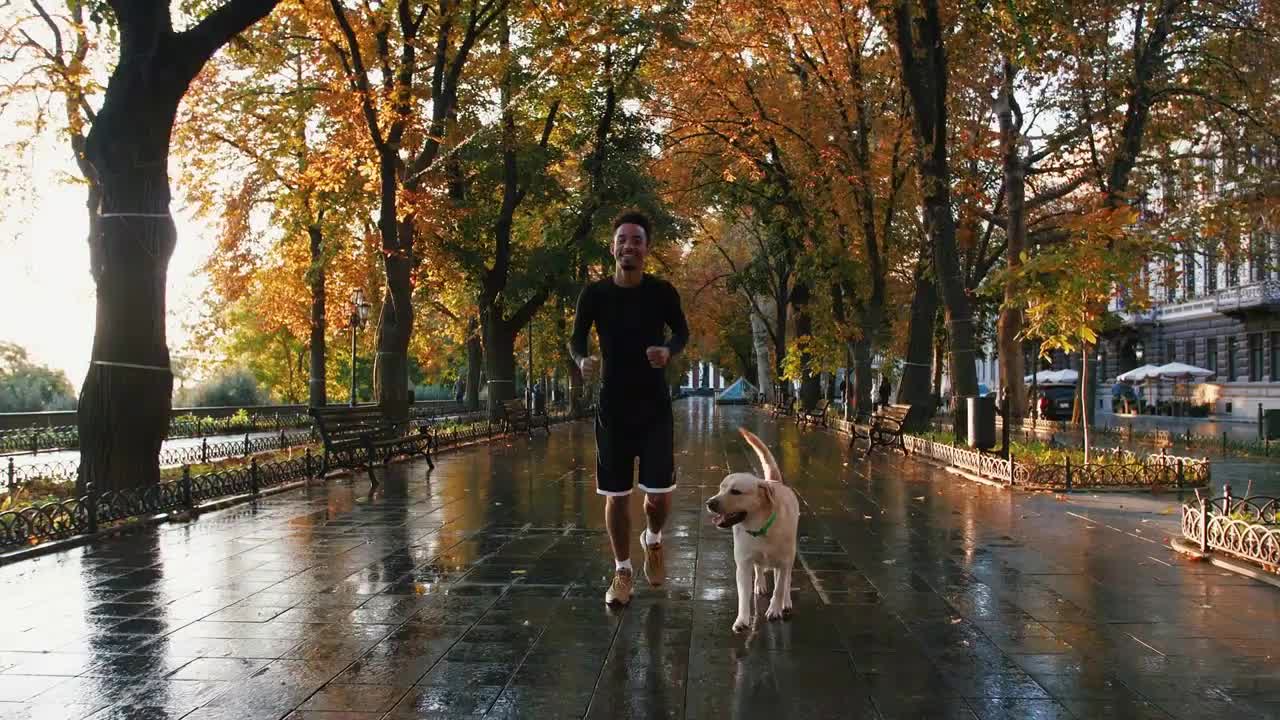 Young black man running with his white labrador dog through the city park during