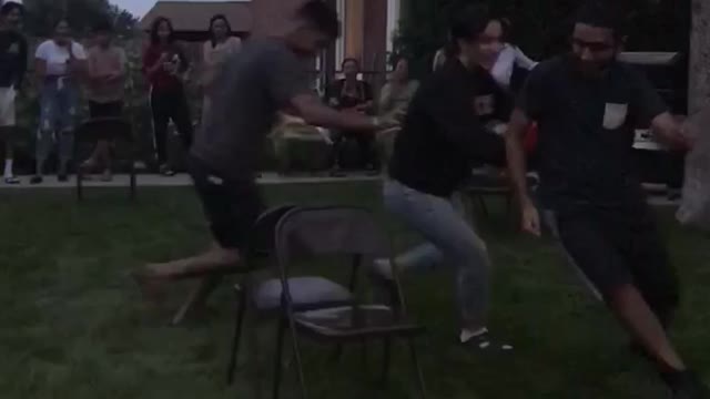 Friends playing musical chairs one friend knocks over a chair and falls in the grass