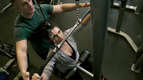 A Man Assisting A Woman In Using The Flat Pulldown Weights Equipment In A Gym