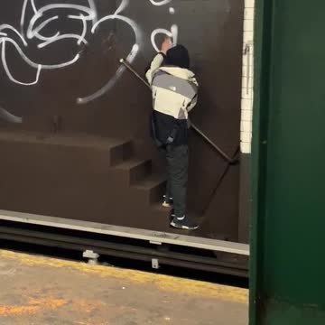 Guy graffiti tags a subway wall behind subway train tracks