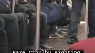 Person riding a crowded subway train wears a bright green cthulhu beanie mask