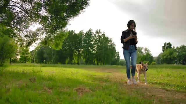 A girl in headphones walks with her dog in the park, listening music from smartphone