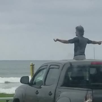 Guy does weird arm warmup while standing out sunroof of truck in front beach