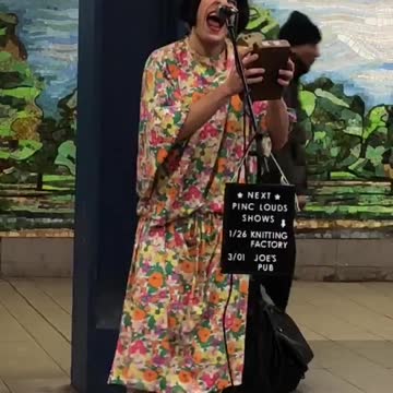 Man in floral dress wig and makeup sings in a subway station