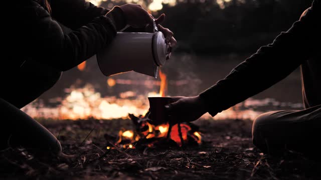 People pouring a warm drink around a campfire