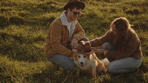 Blonde girl and caucasian young man in hat sitting on grass in field with dog