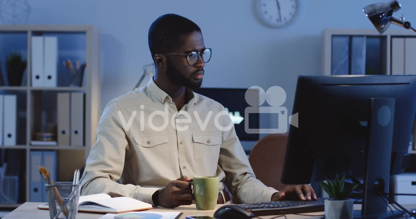 Young Man Drinking Coffee While Working At The Computer In The Office At Night