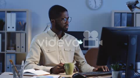 Young Man Drinking Coffee While Working At The Computer In The Office At Night