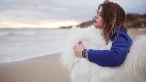 Side view of a young woman sitting on the sand and embracing her dog of the Samoyed breed by the sea