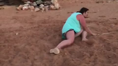 Woman in blue dress attempts to hit golf ball off sand and misses and falls down