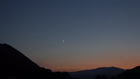 The sun goes down and there's a crescent moon up the mountain.