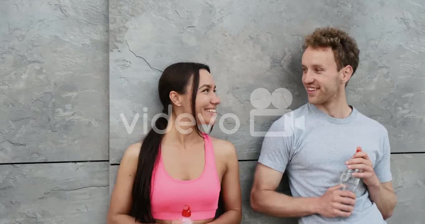 Happy Sportive Couple Leaning Against A Wall, Holding Water Bottle And Talking To Each Other, While