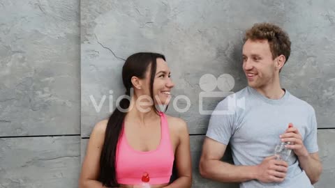 Happy Sportive Couple Leaning Against A Wall, Holding Water Bottle And Talking To Each Other, While