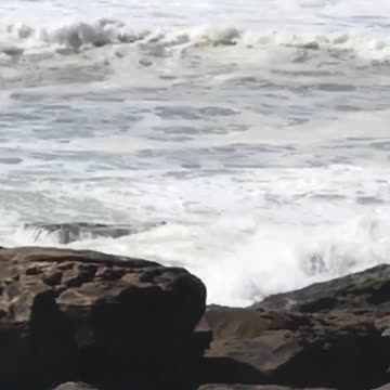 Guy standing on rocks with surfboard gets knocked over by big wave